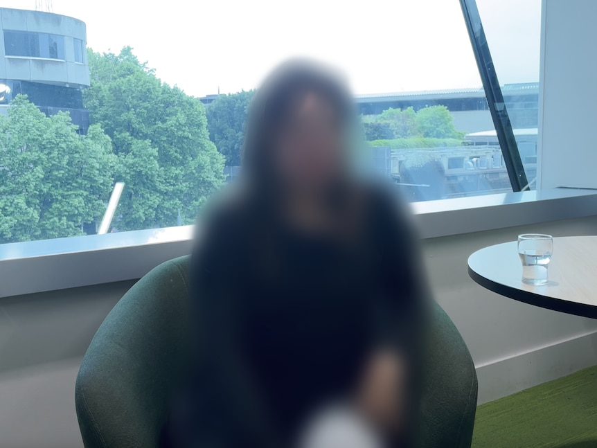 A woman whose face is blurred out sits on a lounge chair in front of a window that looks out over trees