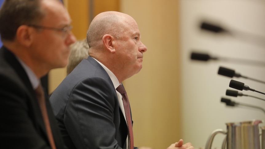 A bald man in a suit and red tie speaks into a microphone while siting at a desk in a room decorated with timber.