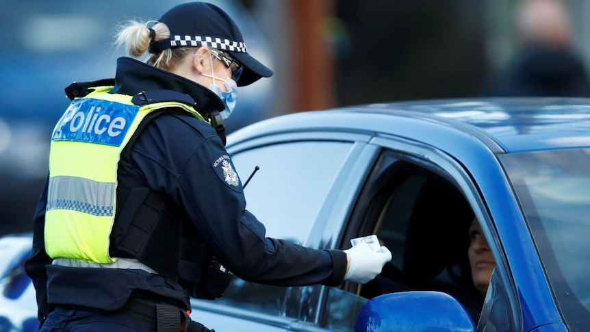 A female police officer with blonde hair wears a facemask and gloves as she checks the licence of a female driver in a blue car.