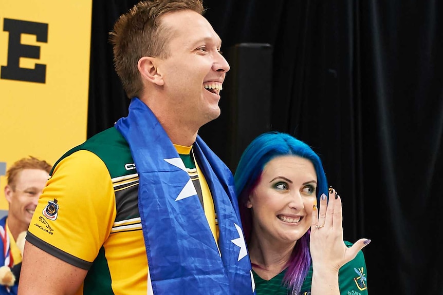 with a backdrop of medal ceremony a man goes down on his knee as a woman holds his face in her hands, bending down in joy