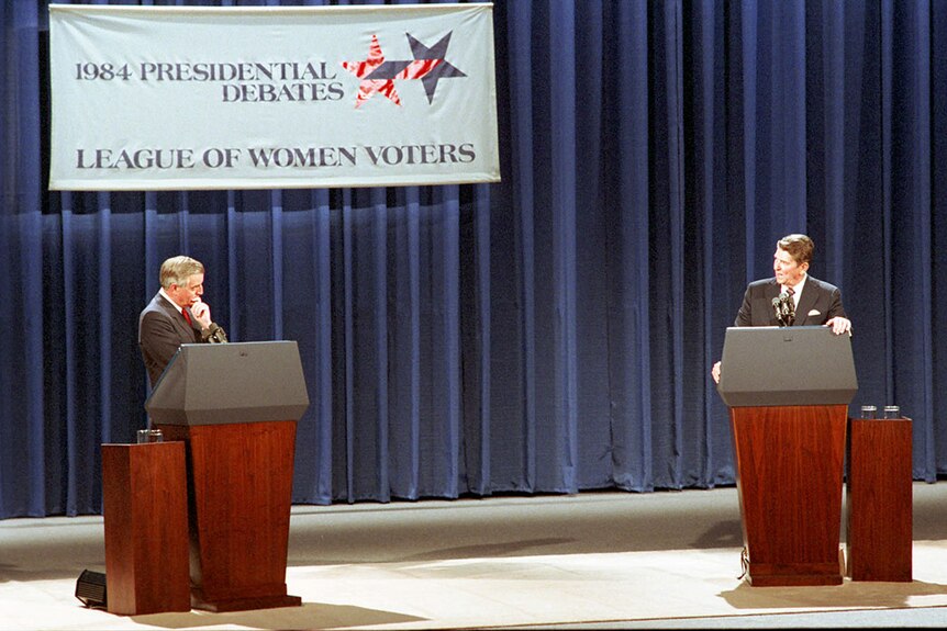 Two older white men in dark suits stand behind wooden lecterns in front of a blue curtain. The man on the right is speaking.