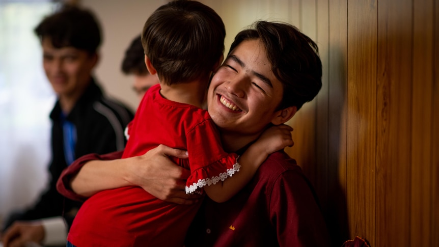 A young man in a maroon top hugs a small child wearing a red shirt