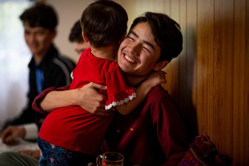 A young man in a maroon top hugs a small child wearing a red shirt