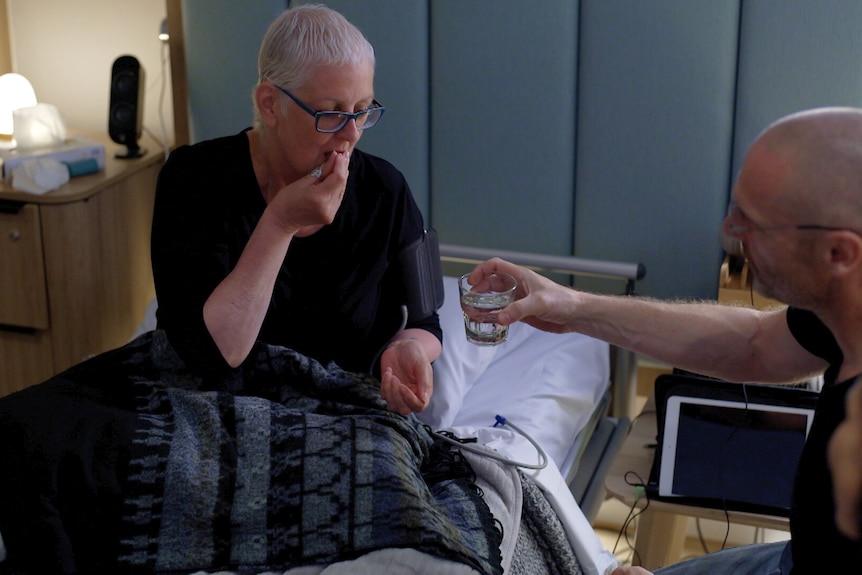 A man hands a woman with short hair and glasses a glass of water as she sits up in bed and swallows a pill