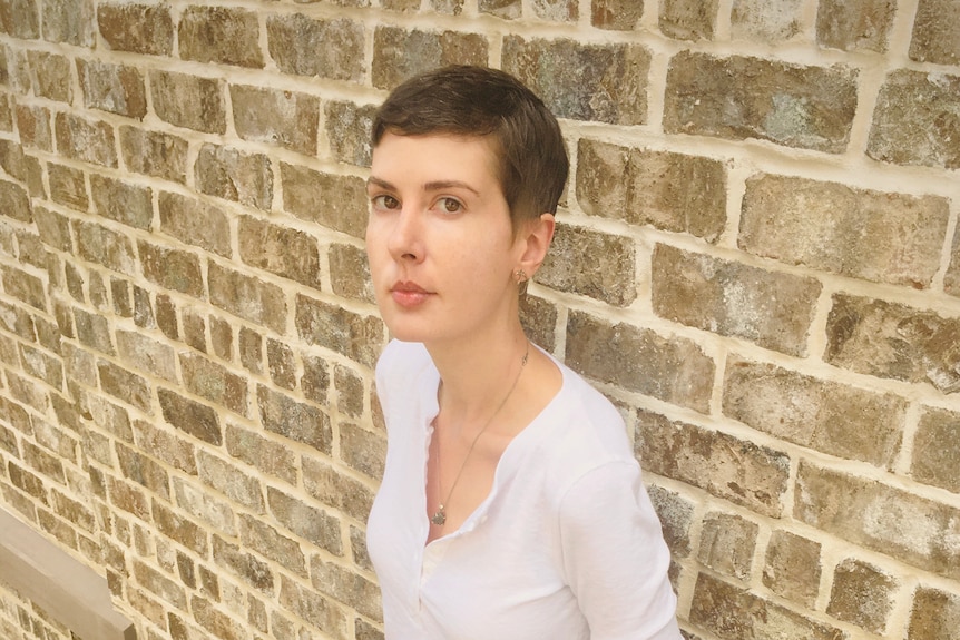 A young white women with cropped brown hair stands leaning against brick wall, looking at camera, wearing white tee.