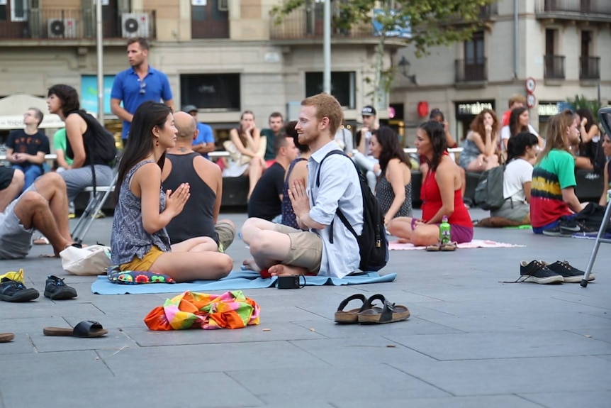 A woman and a man sit cross legged in the CBD facing each other, a crowd of others doing the same.