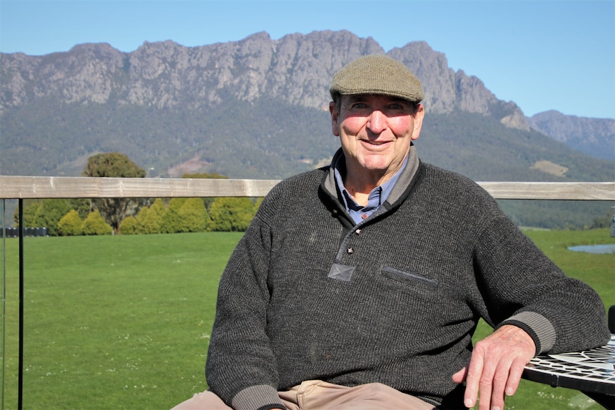 A man wearing a cap sits on a bench with a mountain towering behind him in the distance.