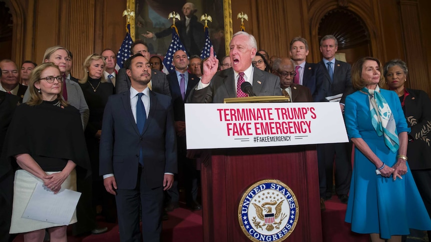 Group of US lawmakers stand as man in centre waves his finger in front of podium with sign 'Terminate Trump's fake emergency'