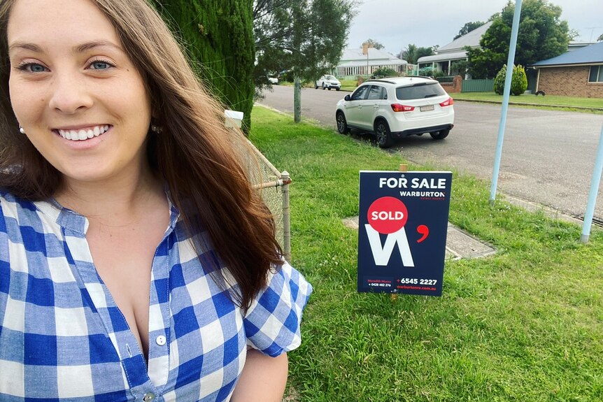 Selfie of a young woman in a checked shirt standing in front of a real estate 'sold' sign.