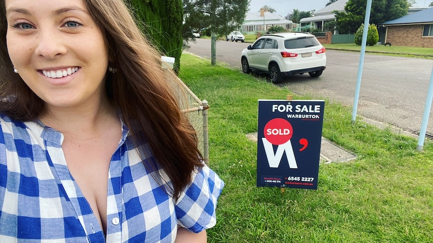 Selfie of a young woman in a checked shirt standing in front of a real estate 'sold' sign.