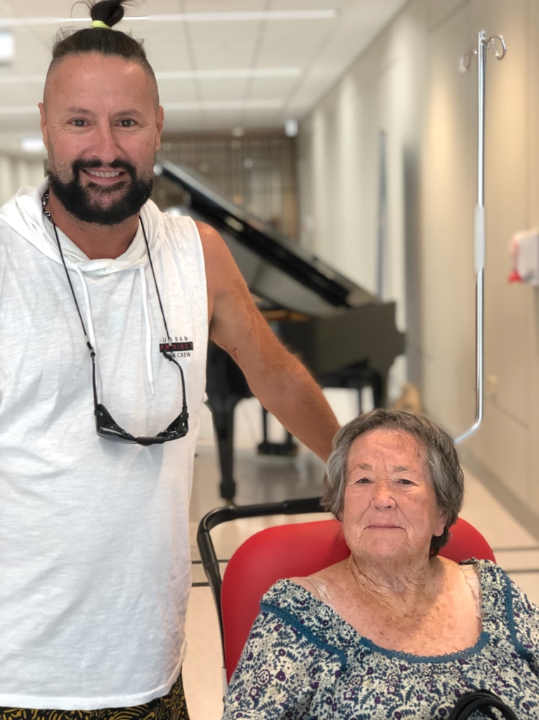 Man stands beside his mum who's in a hospital wheelchair, both smiling.