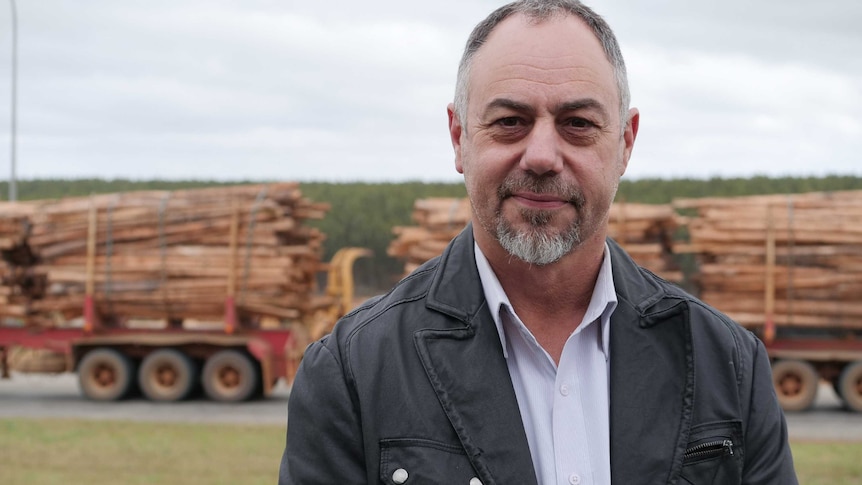 A man with a beard stands in front of a truck carrying timber logs.