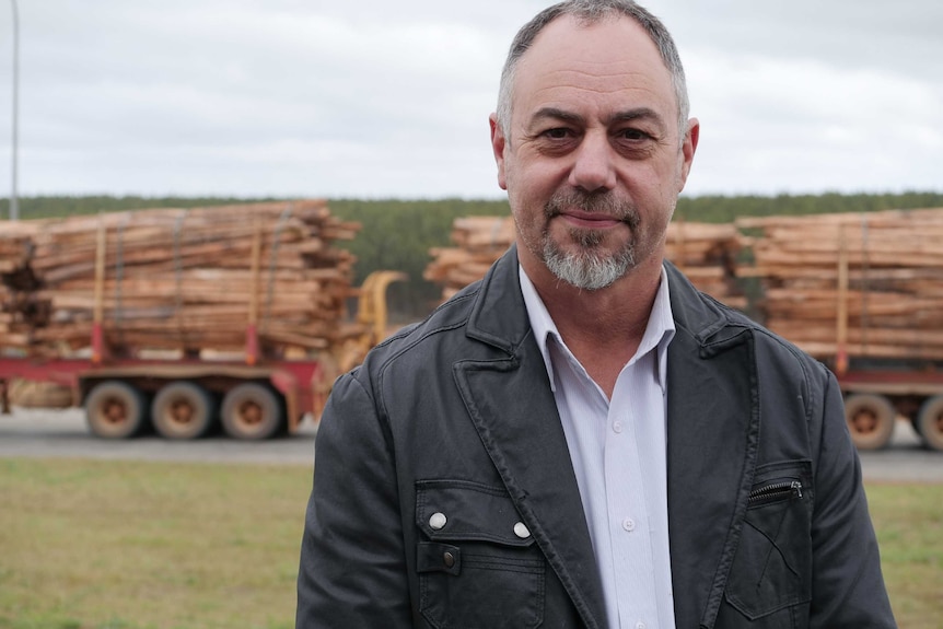 A man with a beard stands in front of a truck carrying timber logs.