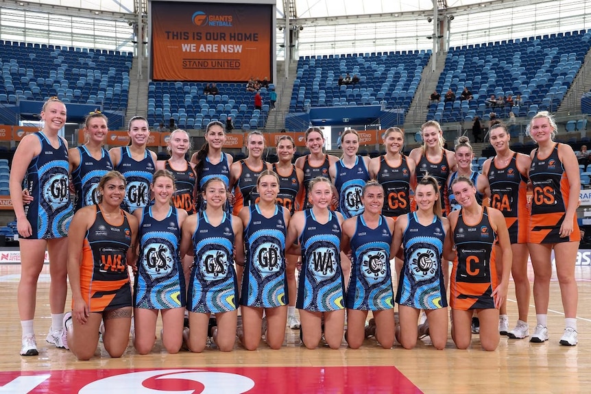 A row of netballers, dressed in uniform, stand arm in arm, while another row of players kneels in front of them.