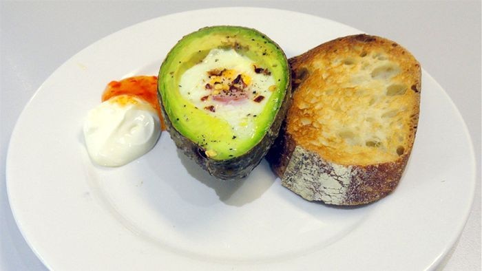 A baked avocado with an egg in the middle where the stone was, with toast, sour cream and sweet chilli sauce, on a plate.
