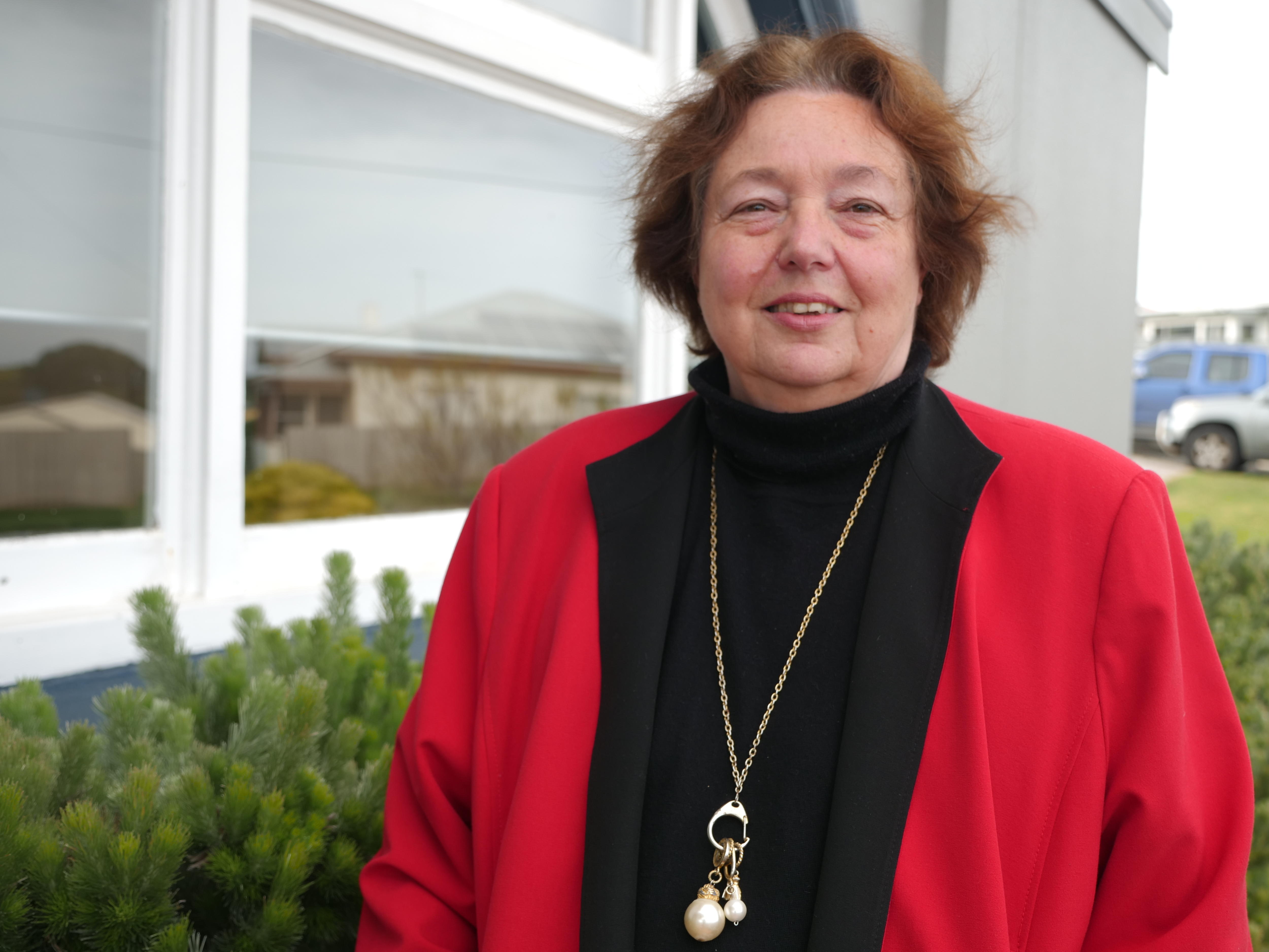 A middle-aged woman standing in front of a window wearing a black shirt and red jacket.