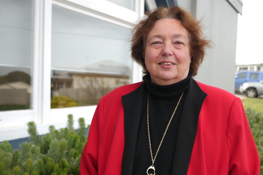 A middle-aged woman standing in front of a window wearing a black shirt and red jacket.