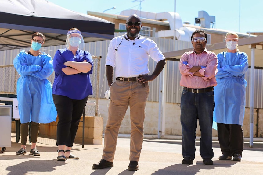 Five medical professionals stand outside, arms crossed, in a V formation, wearing personal protection gear.