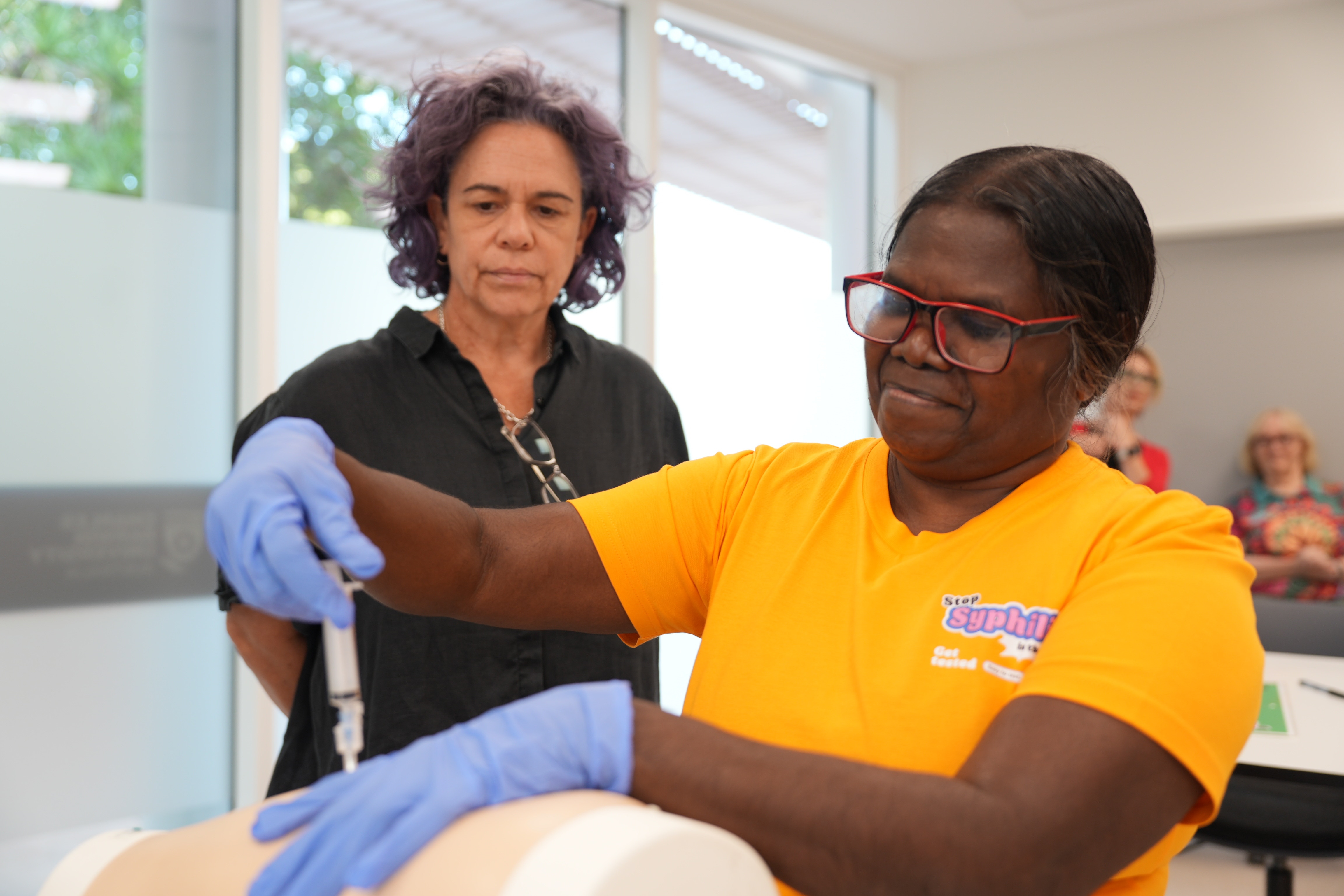 An Indigenous woman injecting a test dummy with a needle, she is wearing glasses and rubber gloves.