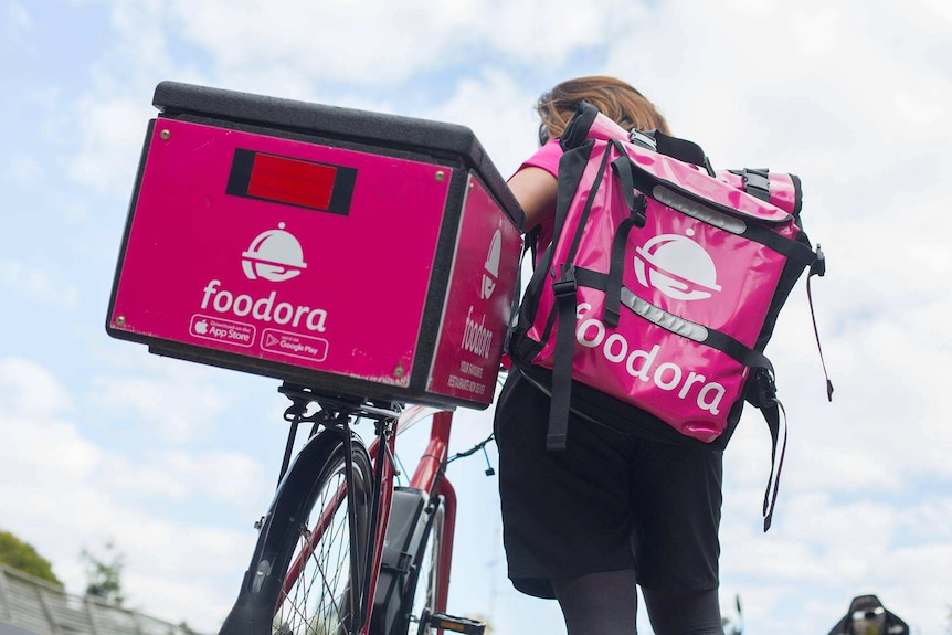 A Foodora delivery worker carries a bag of food and wheels a bicycle.