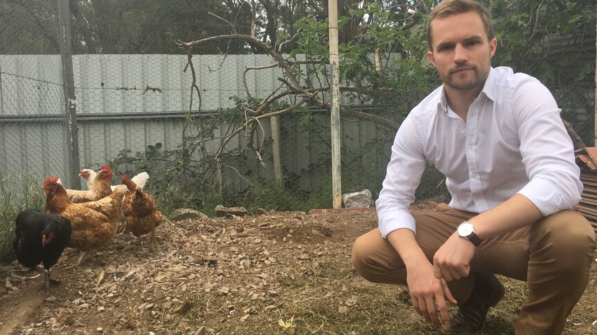 RSPCA senior policy officer Jed Goodfellow crouches in a backyard chicken coup while five hens are grouped behind him.
