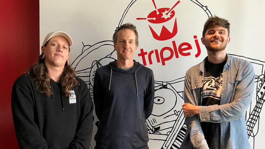 Photograph of Richard Kingsmill standing with two members of Polaris in front of triple j signage.