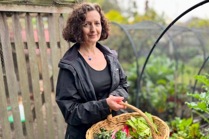 A woman standing in a garden holds a hamper of produce.