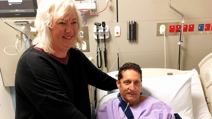 Injured cyclist Peter Duncan lies in a hospital bed with his wife Tanya Duncan standing alongside.