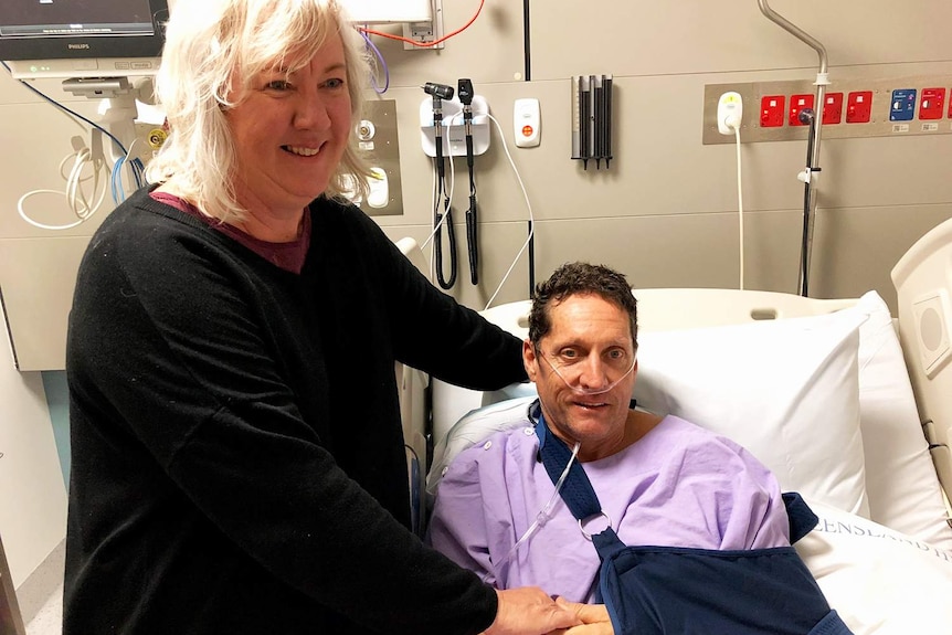Injured cyclist Peter Duncan lies in a hospital bed with his wife Tanya Duncan standing alongside.