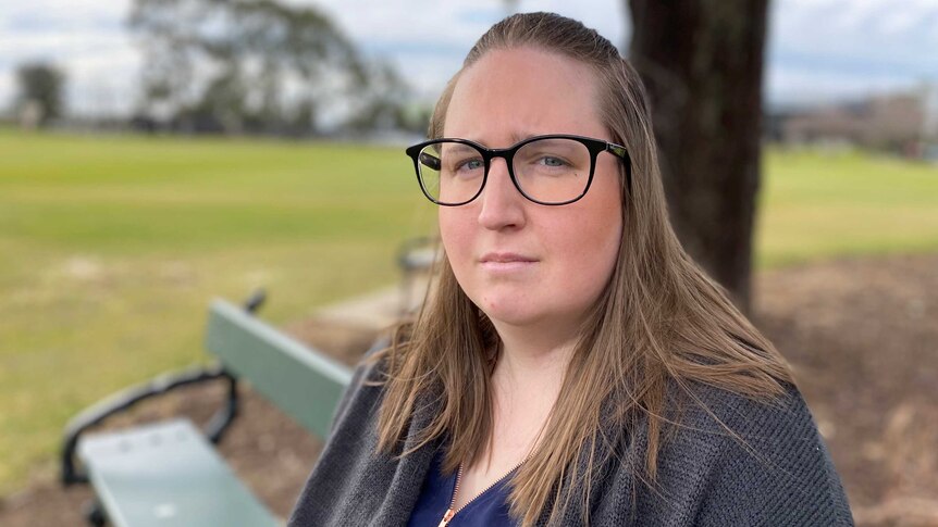 A woman in big glasses looking sombre sitting on a bench looking at the camera