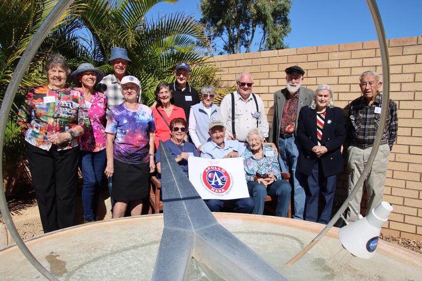 A group of elderly men and women stand in a row behind a steel fountain.