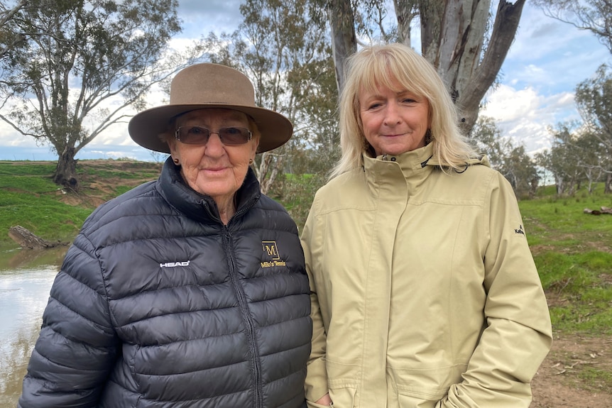 A woman in a black jacket, sunglasses and hat, stands next to a woman in a clay jacket with blonde hair.