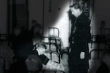 A black and white photo of a Christian Brother standing over a boy next to a bed in a dormitory.