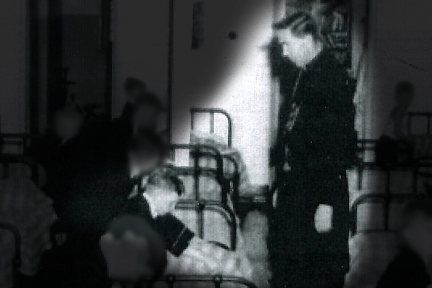 A black and white photo of a Christian Brother standing over a boy next to a bed in a dormitory.