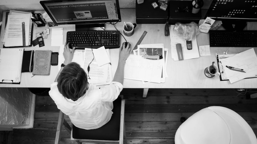 Man works at a crowded desk surrounded by papers and coffee mugs.