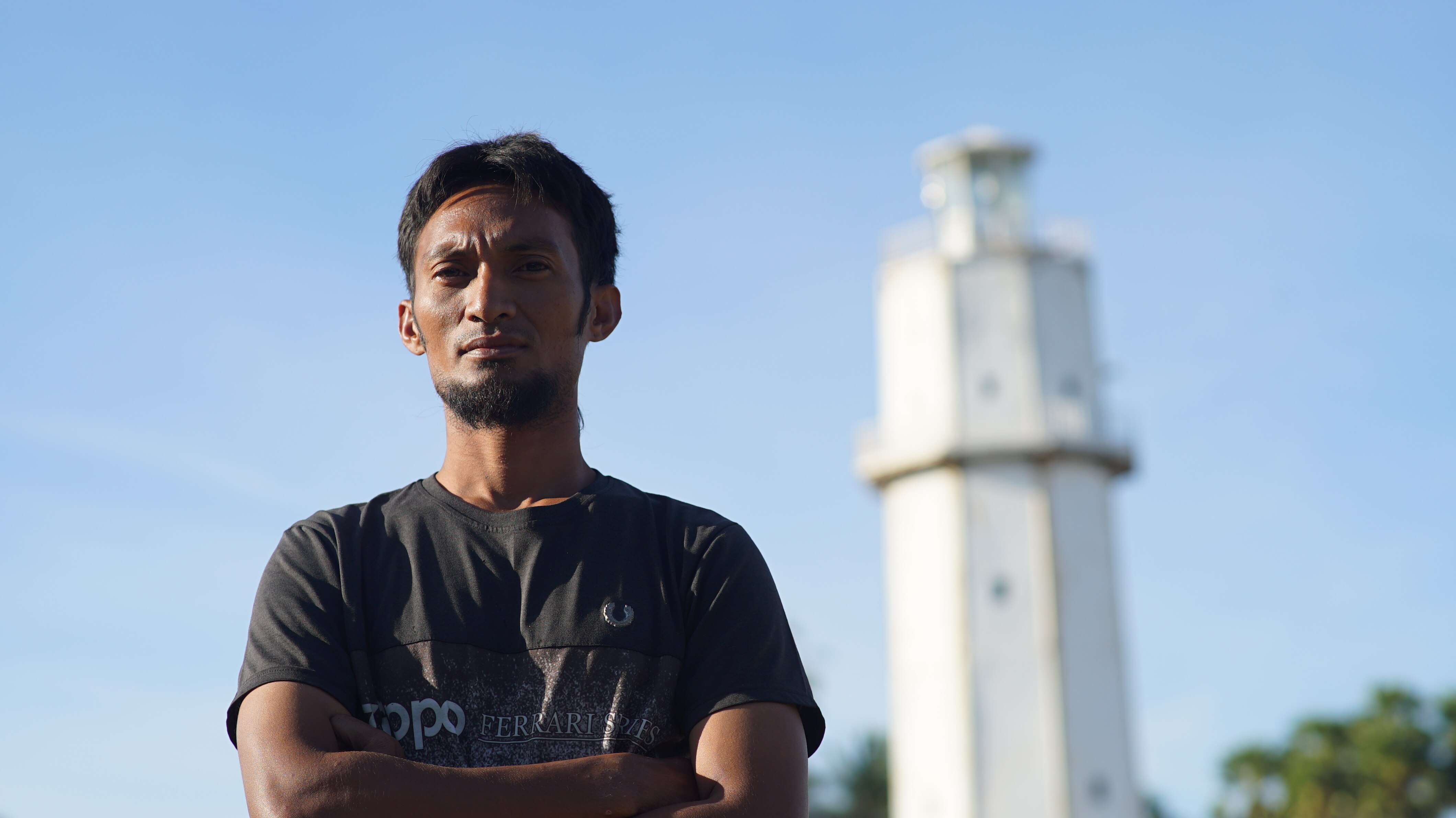 A man standing with arms folded on a blue sky day with a lighthouse in the background