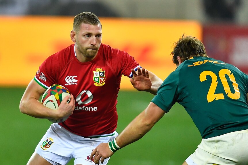 Finn Russell holds a rugby ball under one arm and raises the other to fend off a player in a green shirt