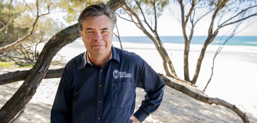A man wearing a navy shirt stands on a sandy beach with trees and the ocean visible in the background.