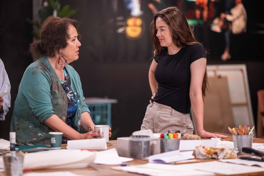 Bronwyn stands at a table, leaning on one arm as she chats to a woman.