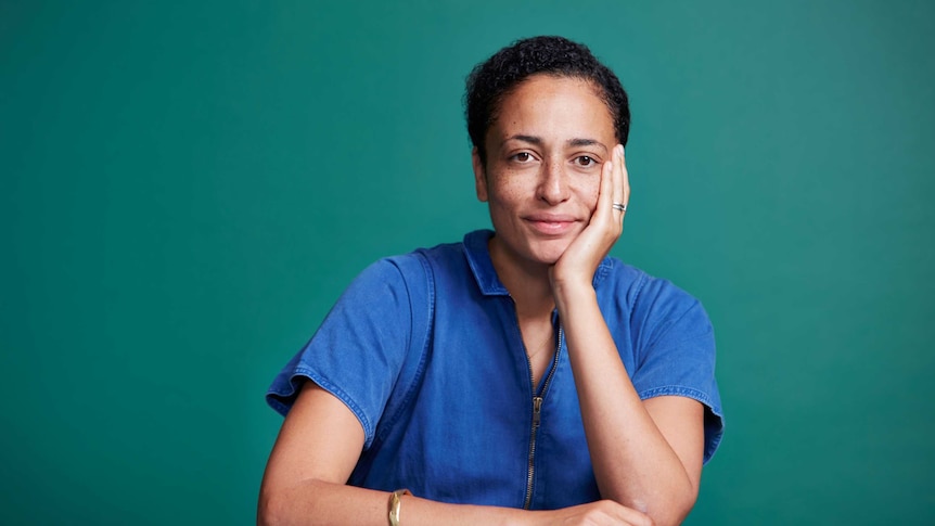 Writer Zadie Smith with one hand holding her face and the other resting on a desk, smiling