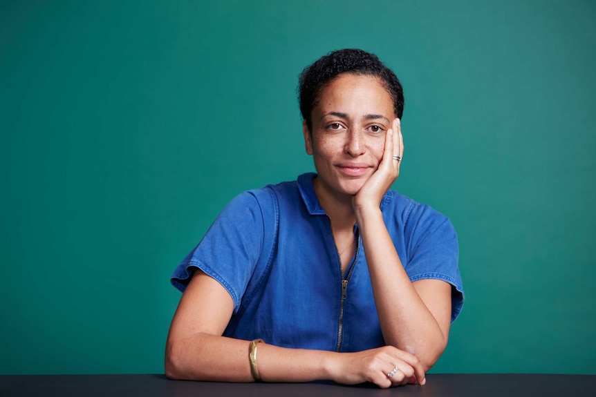 Writer Zadie Smith with one hand holding her face and the other resting on a desk, smiling
