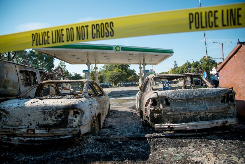 Two burnt cars surrounded by broken glass and debris behind police tape in Milwaukee BP parking lot.