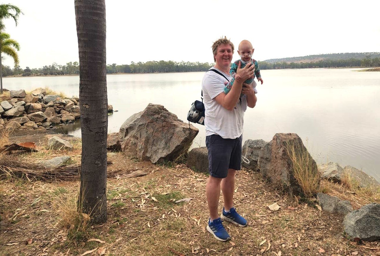 A man stands on the water's edge next to a palm tree holding his 13 month old son.