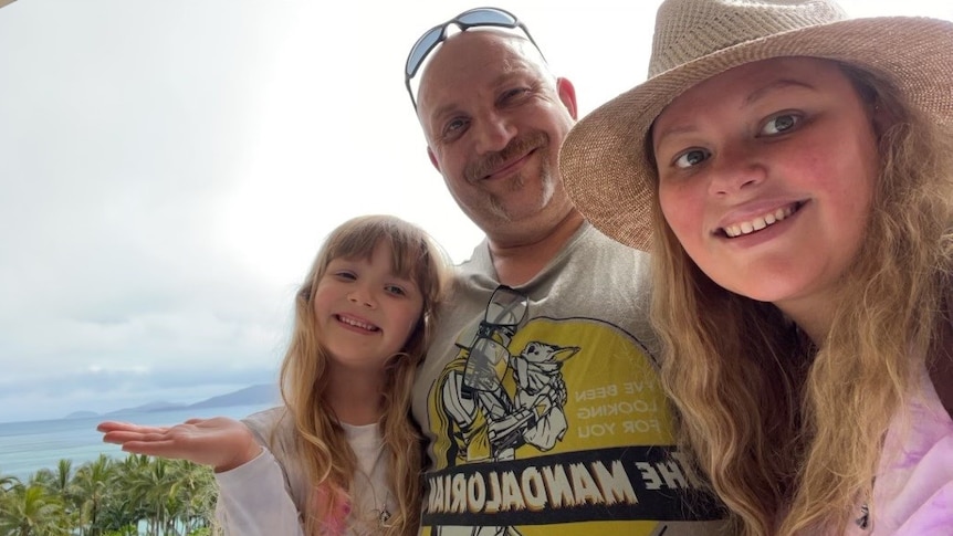 A woman, a man and a young girl huddle together smiling at the camera with palm trees and ocean behind them.