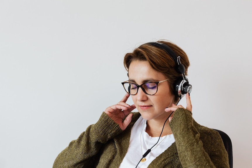 A person with short hair is seen wearing a khaki cardigan while holding their hands over their headphones, eyes closed