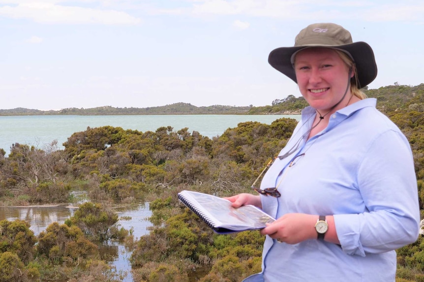 Woman in hat on right with notebook standing in front of saltmarsh area in front of ocean inlet