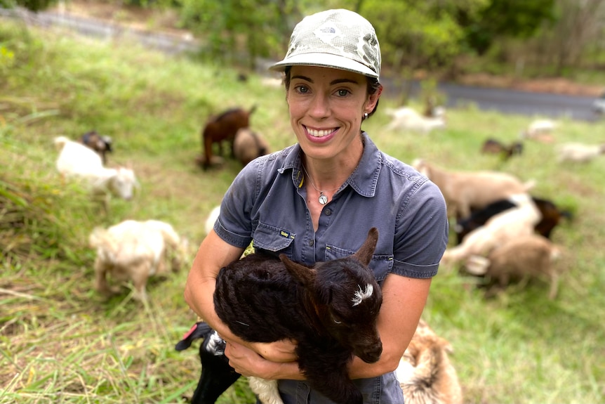 A woman wearing a hat is smiling while holding a baby goat on a steep slope