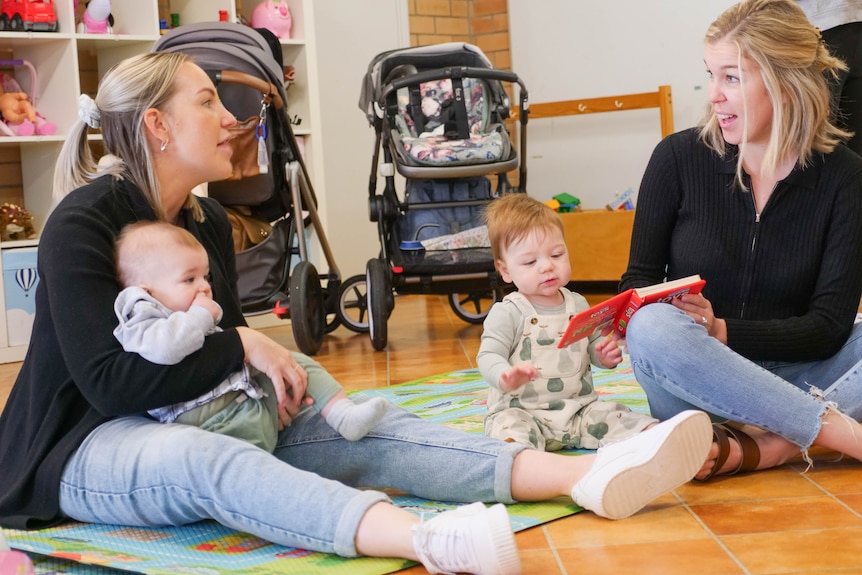 Two mothers sitting on the floor with babies next to them chatting. 