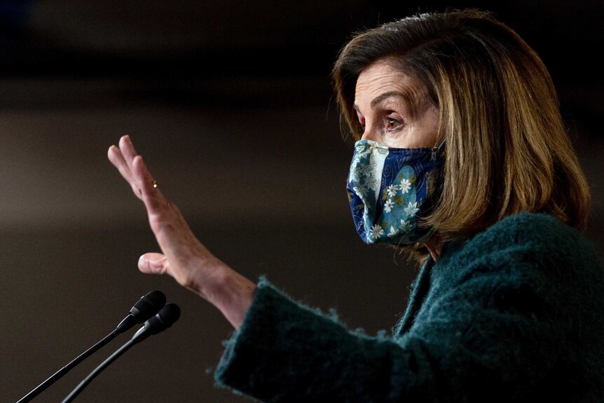Wearing a floral face mask, Nancy Pelosi gestures with her right hand while speaking at a lectern