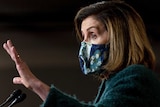 Wearing a floral face mask, Nancy Pelosi gestures with her right hand while speaking at a lectern